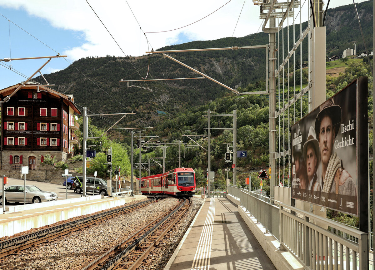 An der Matterhorn-Gotthard Bahn MGB: Einfahrt in den Bahnhof Stalden. Ein Zug mit  Komet  Triebzügen, geführt vom Doppelsteuerwagen ABt 2131. Rechts Werbung für das Drama  Iischi Gschicht  (Unsere Geschichte), die von der (in eine Katastrophe mündenden) Erstbesteigung des Matterhorns 1865 handelt. 30.August 2025 