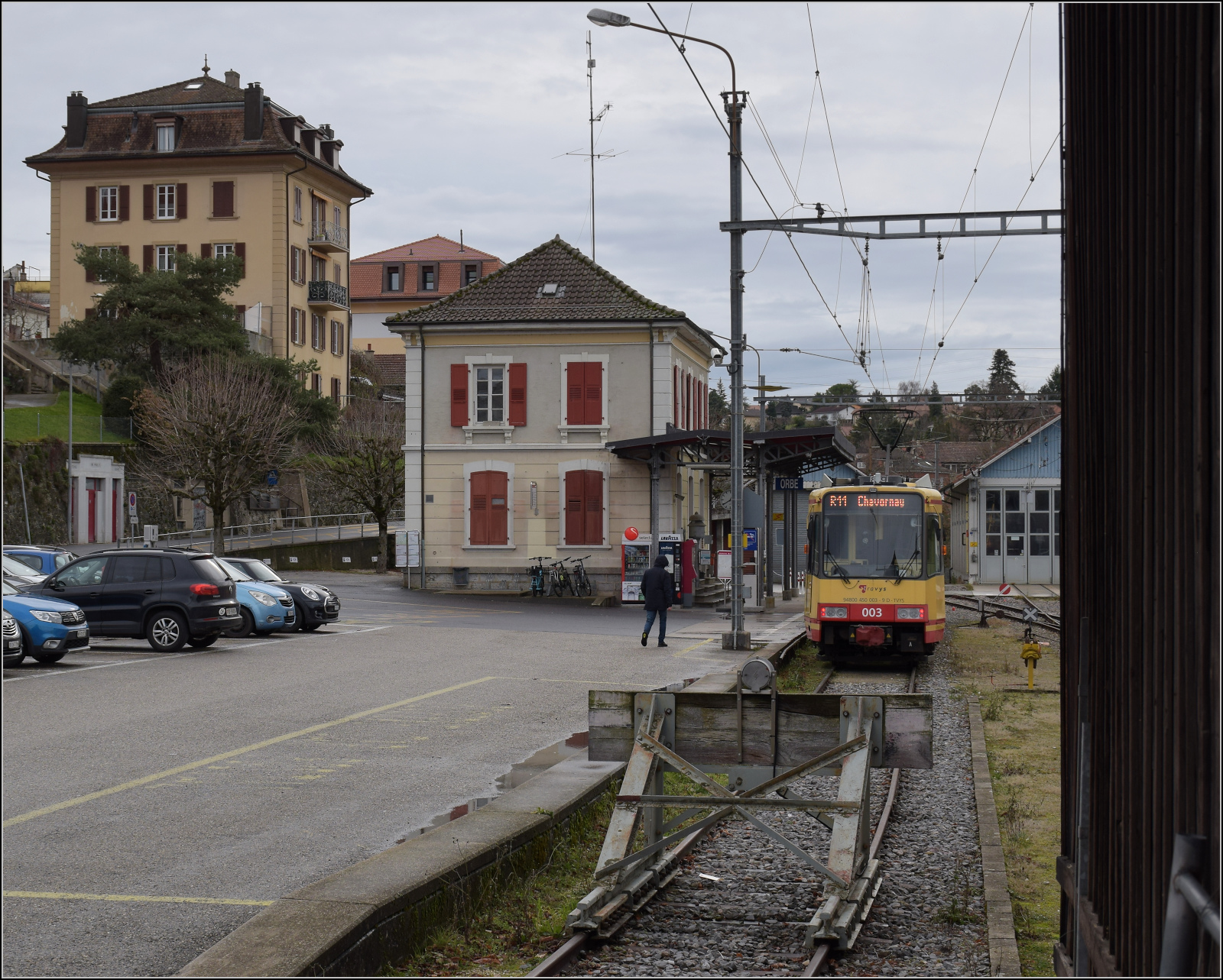 Ende einer Ära oder Gleichstrombetrieb adieu.

Die Kleinstadt Orbe teilt sich ein Problem vieler Orte entlang früh gebauter Bahnlinien. Bahnstrecken mussten wegen mangelnder Zugkraft der frühen Lokomotiven möglichst in der Ebene gebaut werden. Zudem waren anfänglich die Belange der Fahrgäste eher zweitrangig, Bahnen bauten passend für ihren Betrieb. Viele historische Städte waren aber auf strategisch gut zu verteidigenden Anhöhen gebaut und wurden von den Bahnstrecken umfahren. Somit drohte all diesen Orten ein wirtschaftlicher Abstieg, da die Bahn für die Logistik immer wichtiger wurde. Ziehmlich häufig traf es Orte in Bayern, wo die Bahnverwaltung aus heutiger Sicht eher selbstzentriert arbeitete. Ein Lösung brachte das Lokalbahngesetz, das somit auch eine neue Klasse Eisenbahnen schuf, womit die Erschließung je nach lokaler Initiative von kleinen Unternehmen schnell erfolgte. Orbe dürfte nebst Balsthal das wohl prominenteste Beispiel der Schweiz mit einer parallelen Entwicklung zur  Lokalbahn  sein.

Da das Elektrizitätswerk Orbe Betreiber der Bahn werden sollte, lag es nahe, die seinerzeit 'hypermoderne' Elektrizität auch gleich für die neueste Entwicklung der Bahnwelt zu nutzen, die elektrische Eisenbahn. So entstand 1894 womöglich die erste elektrische Normalspurbahn auf dem europäischen Kontinent, noch ein Jahr vor der ersten deutschen Normalspur-Strecke Tettnang-Meckenbeuren. Die Inbetriebnahme fand auch nur ein Jahr nach der normalspurigen Liverpool Overhead Railway statt. Zu dieser Zeit steckte die Technik noch in den Kinderschuhen, so dass mit 700 Volt eine bescheidene Spannung genutzt wird. Zwar sind zwei wichtige Fahrzeuge im Museum gelandet, dennoch überrascht dieser sang- und klanglose Abschied von dieser Technik, deren Implementierung man eigentlich einst als sensationell bezeichnen könnte. Deswegen ist zum endgültigen Ende des Gleichstrombetriebs nur noch ein Gastarbeiter aus dem grossen Kanton auf der Strecke mit Gleichstrom unterwegs. Für authentischen Betrieb aus diesen Anfangstagen muss man jetzt nach Trossingen in Baden-Württemberg fahren, wo mit Zeug Christe ein Triebwagen aus dem Jahr 1898 an ganz wenigen Fahrtagen unterwegs ist.

Der Karlsruher Zweisystem-Strassenbahn  Be 4/8 003  alias 94 800 450 003-9 D-TVYS wartet im Bahnhof Orbe auf seine nächste Runde, um die Fahrgäste zu den S-Bahnen in Chavornay zu bringen. Dezember 2025.

Und in diesem Sinne werden wir sehen, wie der neue Service nach dem direkten Anschluss an Lausanne angenommen wird, wenn die Leitungen und die neue Verbindungskurve zum Bahnsteig in Chavornay in Betrieb sein wird.
