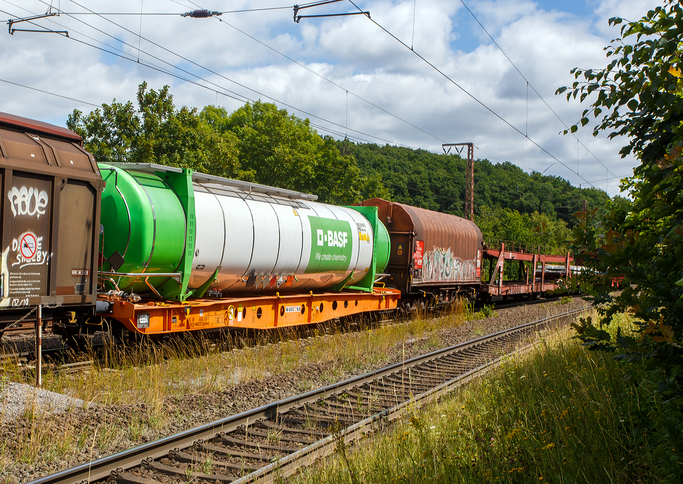 Vierachsiger Containertragwagen 33 85 4505 278-3 CH-WASCO der Gattung Sgmmns 54‘, für den Transport von intermodalen Ladeeinheiten und des Wascosa flex freight system (Wechselaufbauten), der Wascosa AG am 11 Juli 2025 im Zugverbund bei einer Zugdurchfahrt in Rudersdorf (Kreis Siegen). Der Wagen ist beladen, mit einem vom belgischen Hersteller Van Holl gebauten, 52 ft Tankcontainer der BASF, der hier laut Gefahrguttafel 83/2734 mit Amine oder Polyamine befüllt ist. Die Kombination des Wagens mit 45‘ oder 52‘ Tankcontainern kann Kesselwagenersetzen und ermöglicht eine unabhängige Instandhaltung beider Komponenten.

Gebaut wurde der Wagen vom slowakischen Güterwagenhersteller Tatravagónka in Poprad.

TECHNISCHE DATEN (Wagen):
Spurweite: 1.435 mm (Normalspur)
Länge über Puffer: 17.800 mm
Drehzapfenabstand: 11.920 mm
Radsatzstand in den Drehgestellen: 1.800 mm
Ladelänge: 16.470 mm
Höhe der Ladeebene für Container über S.O.: 1.105 mm
Drehgestell: Y25 Lsi(f)-C-K
Raddurchmesser: 920 mm (neu) / 860 mm (abgenutzt
Eigengewicht: 16.500 kg
Max. Zuladung bei Lastgrenze S: 73,5 t (ab Streckenklasse D)
Max. Geschwindigkeit: 100 km/h (leer 120 km/h)
Max. Radsatzlast: 22,5 t
Druckluftbremse: Knorr KE-GP-A (K)
Bremssohle: C 810
Handbremse: Ja
Kleinster bef. Gleisbogenradius: 150 m (im Zugverband) / 75 m der Einzelwagen
Intern. Verwendungsfähigkeit: TEN GE

Der Tankcontainer:
Der 52-Fuß-Tankcontainer mit 75 Tonnen zulässigem Gewicht (bei einem Eigengewicht von 8.200 kg) und 73.500 Liter Inhalt, hat die doppelte Ladekapazität wie ein heute üblicher Tankcontainer und eine vergleichbare Kapazität wie ein Chemiekesselwagen. Der neue, bahnoptimierte Tankcontainer hat die gleiche Technik wie die bisher üblichen 20 bis 30 Fuß messenden Tankcontainer. Die 52 Fuß-Tankcontainer können zum Bahntransport, Binnenschiffstransport und, im leeren Zustand, auch zum Straßentransport eingesetzt werden. Darüber hinaus sind sie zur Lagerung in Containerlagern zugelassen. Ausgestattet sind sie außerdem mit moderner Isoliertechnik sowie Heizvorrichtungen. Der 52 Fuß-Tankcontainer verfügt im Vergleich zum Bahnkesselwagen über ein höheres Ladegewicht im Verhältnis zum Eigengewicht. Dank der Möglichkeit, vom Bahnwagen abgenommen werden zu können, und der Stapelbarkeit benutzt der Tankcontainer deutlich weniger Infrastruktur und ist flexibler beim Be- und Entladen.

Entwickelt wurden die 45 und 52 Fuß Tankcontainer für den Bahnverkehr von Van Hool und BASF gemeinsam. Der erste Prototyp wurde 2015 geliefert.